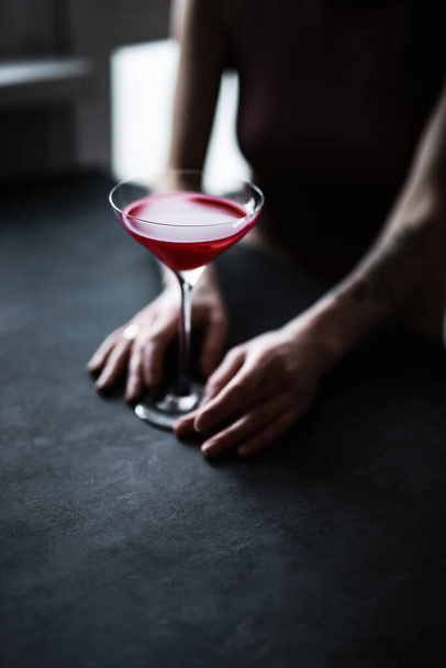 Ένα κόκκινο βατόμουρο ξινό κοκτέιλ σε ένα όμορφο ποτήρι κουπέ στα χέρια του κοριτσιού, πίσω φως, σκούρο και κυκλοθυμική φωτογραφία - Φωτογραφία, εικόνα