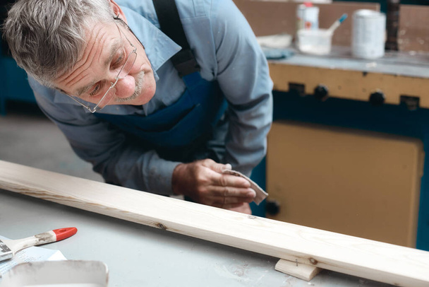 Οι ξυλουργοί δουλεύουν στο ξυλουργείο. Ένας ηλικιωμένος αρσενικός καμπινετίστας εξετάζει προσεκτικά μια ξύλινη σανίδα.  - Φωτογραφία, εικόνα