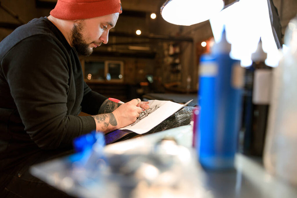 Profesional hombre barbudo caucásico tatuaje maestro está trabajando en acogedor estudio atmosférico - Foto, Imagen