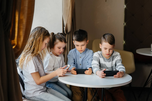 Children sit at a table in a cafe and play mobile phones together. Modern entertainment - Фото, изображение
