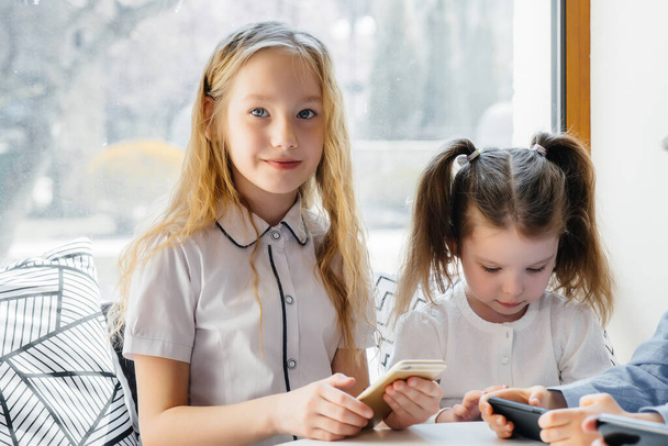 Children sit at a table in a cafe and play mobile phones together. Modern entertainment - Valokuva, kuva