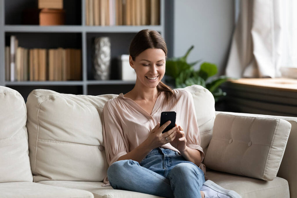 Smiling woman looking at smartphone screen, sitting on couch - Fotografie, Obrázek