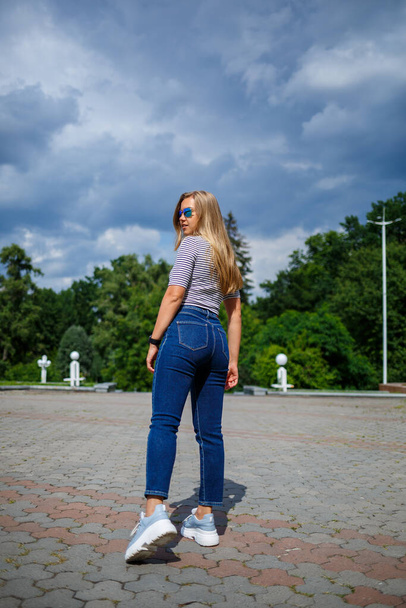 Una hermosa chica con el pelo largo y rubio de aspecto europeo. Vestido con jeans y una camiseta. Caminar en un día soleado - Foto, Imagen