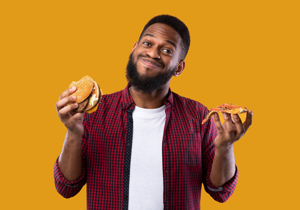 Africký muž drží Burger a plátek pizzy, žluté pozadí - Fotografie, Obrázek