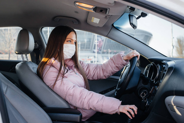 A young girl sits behind the wheel in the car in the mask during the global pandemic and coronavirus. Quarantine - Zdjęcie, obraz