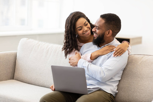 Afrikanisches Paar sitzt auf Couch, benutzt Laptop, kuschelt - Foto, Bild