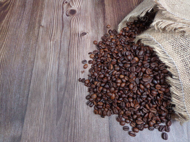 Granos de café tostados apilados saliendo de un saco de tela sobre una mesa de madera, con espacio para colocar texto - Foto, Imagen