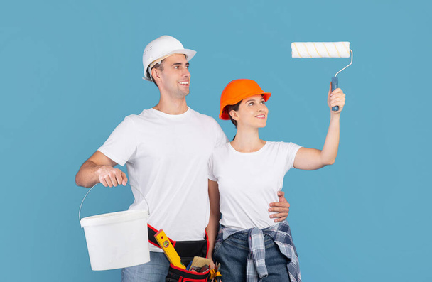 Servicios de renovación de casas. feliz pareja en Hardhats sosteniendo pintura rodillo y cubo - Foto, imagen