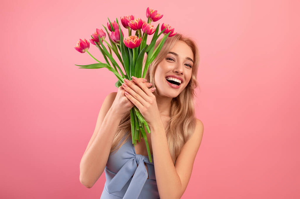 Happy Womans Day. Excited young lady in cocktail dress holding bouquet of tulips on pink studio background - Photo, Image