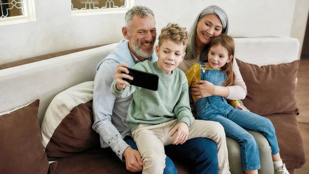 Happy grandparent with two kids grandchildren making a selfie on smartphone while relaxing together on sofa in the living room, hugging and having fun - 写真・画像