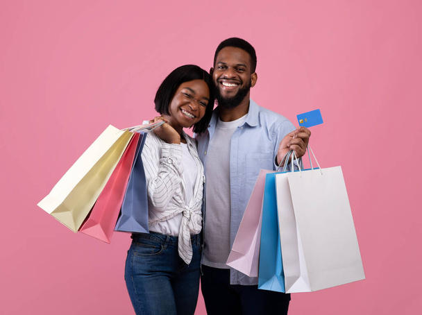 Positieve Afro-Amerikaanse paar met credit card en boodschappentassen glimlachen en poseren op roze studio achtergrond - Foto, afbeelding