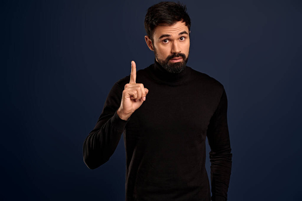 Handsome bearded man in black high neck sweater raise one finger and sharing his thought, suggest something during office meeting, give advice or recommend product, standing blue background. - Fotografie, Obrázek