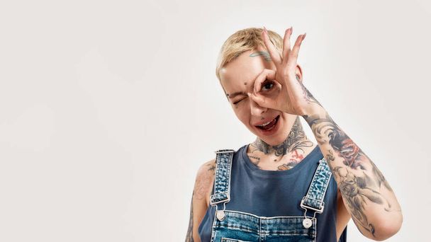 Emotions and postures. A white pierced and tattooed woman showing an OK sign in front of her eye with her mouth open smiling and looking into a camera wearing a denim overall - Photo, Image