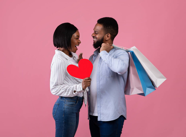 Shopping für den Valentinstag. Schwarzes Paar mit roten Papierherzen und Geschenktüten, die sich auf rosa Hintergrund anschauen - Foto, Bild
