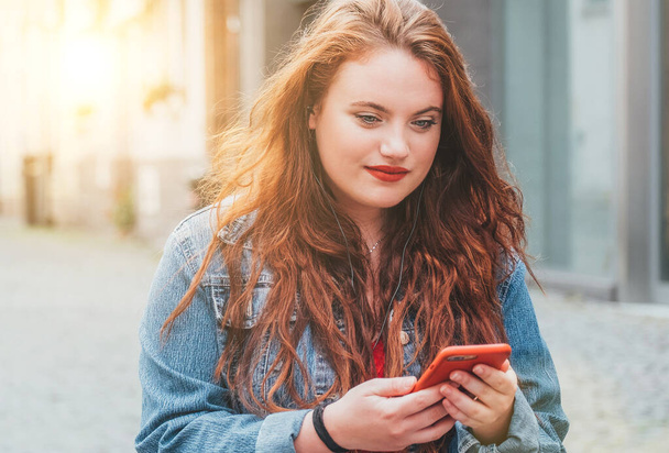 Portrait of red curled long hair caucasian teen girl walking on the street and browsing an internet using a modern smartphone. Modern people with technology devices concept image. - Foto, immagini
