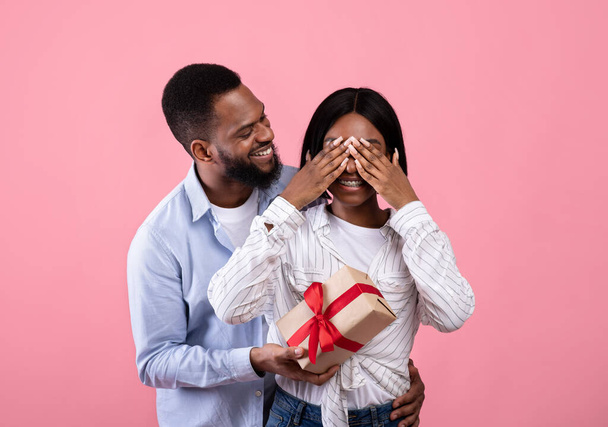 Fürsorglicher afroamerikanischer Mann bedeckt die Augen seiner Freundinnen und überrascht sie mit einem Geschenk auf rosa Hintergrund - Foto, Bild