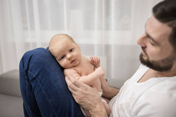 Любящий счастливый отец с бородой держит на руках новорожденного ребенка. Сидит дома на диване. Концепция воспитания, отцовства, первые месяцы жизни ребенка. - Фото, изображение