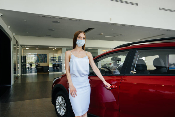 A young pretty girl inspects a new car at a car dealership in a mask during the pandemic. The sale and purchase of cars, in the period of pandemia - Photo, image