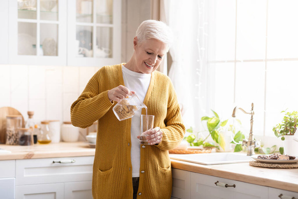 Atractiva abuela vertiendo agua en el vidrio en la cocina en casa - Foto, imagen