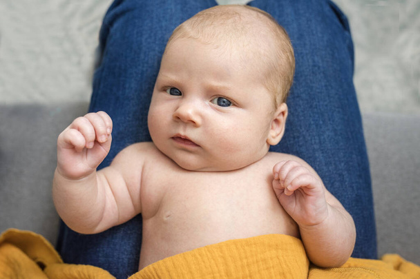 Портрет новорожденного ребенка, лежащего на ногах у родителя дома. Милый голый ребенок, белый, зрительный контакт. Стиль жизни. Модный жёлтый муслиновый подгузник. Концепция современного воспитания, красивые вещи для новорожденного - Фото, изображение