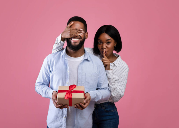 Valentinsüberraschung. Schöne schwarze Frau bedeckt die Augen ihrer Freunde und macht HUSH-Geste auf rosa Hintergrund - Foto, Bild
