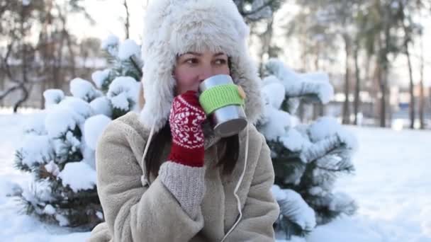 Una mujer joven se para en un bosque o parque invernal y bebe té caliente - Metraje, vídeo