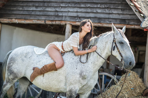 Beautiful brunette woman with long hair and long white dress riding on a white horse. Sensual and attractive  girl with brown boots interacting and having fun with a horse at the ranch - Zdjęcie, obraz