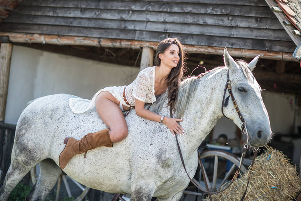 Beautiful brunette woman with long hair and long white dress riding on a white horse. Sensual and attractive  girl with brown boots interacting and having fun with a horse at the ranch - Zdjęcie, obraz