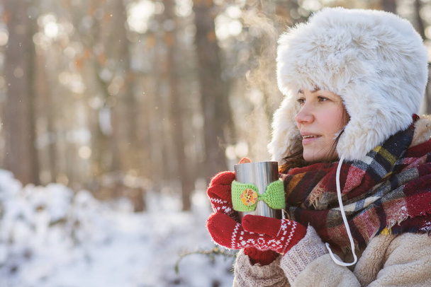 Una mujer joven se para en un bosque o parque invernal y bebe té caliente. - Foto, imagen