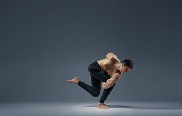 Male yoga keeps balanc in a difficult pose, grey background. Strong man doing yogi exercise, asana training, top concentration, healthy lifestyle - Photo, Image