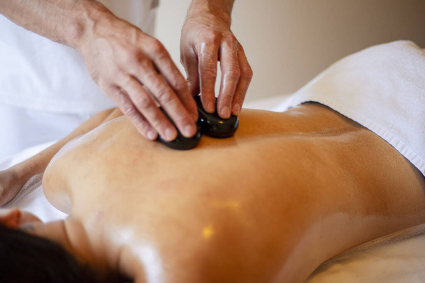 Hot-Stone-Massage, Steintherapie, Thai-Massage, Entspannungstreff, Heilverfahren, ein Mädchen mit nacktem Rücken liegt, schwarze Steine in den Händen - Foto, Bild