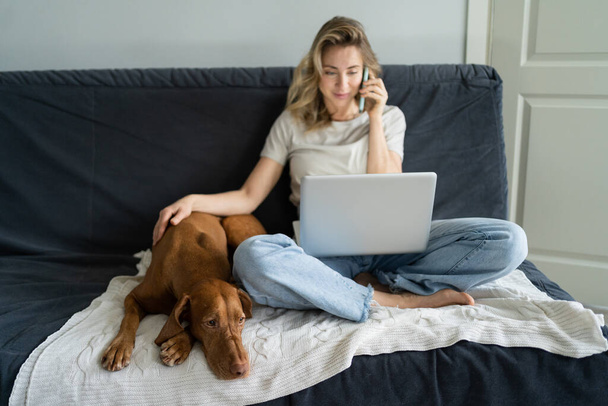 Женщина сидит на диване с собакой, разговаривает по мобильному телефону, дистанционно работает на компьютере ноутбука дома  - Фото, изображение