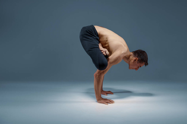 El yoga muscular mantiene el balanceo en las manos en posición difícil, posición de meditación, fondo gris. Hombre fuerte haciendo ejercicio yogui, entrenamiento asana, concentración superior, estilo de vida saludable - Foto, Imagen