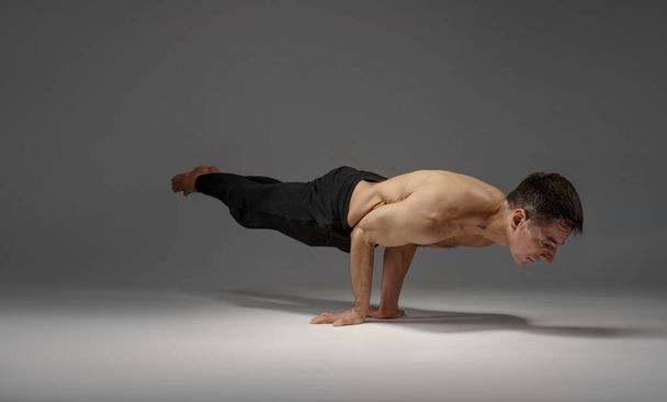 Male yoga keeps horizontal balanc on hands, meditation position, grey background. Strong man doing yogi exercise, asana training, top concentration, healthy lifestyle - Photo, Image