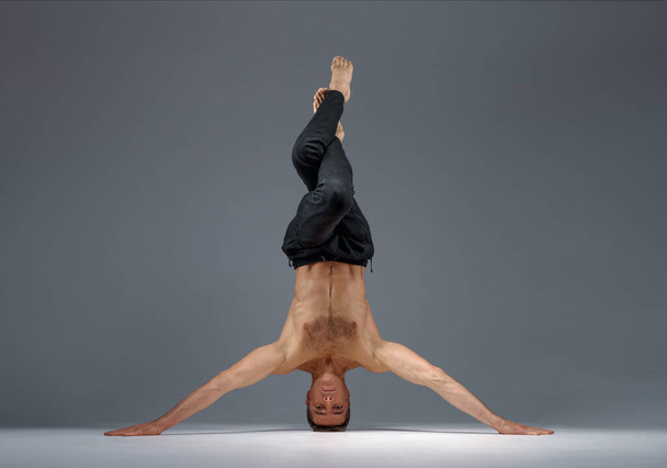 Male yoga standing on his head and hands, meditation, grey background. Strong man doing yogi exercise, asana training, top concentration, healthy lifestyle - Photo, Image
