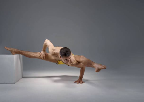 El yoga muscular mantiene al balancín en una postura difícil en el pedestal, posición de meditación, fondo gris. Hombre fuerte haciendo ejercicio yogui, entrenamiento asana, concentración superior, estilo de vida saludable - Foto, imagen