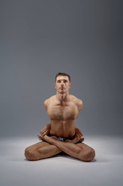 Йога сидит в классической позе, в положении посредника, идеально растягиваясь, на сером фоне. Сильный человек делает йоги упражнения, асана тренировки, высокая концентрация, здоровый образ жизни - Фото, изображение