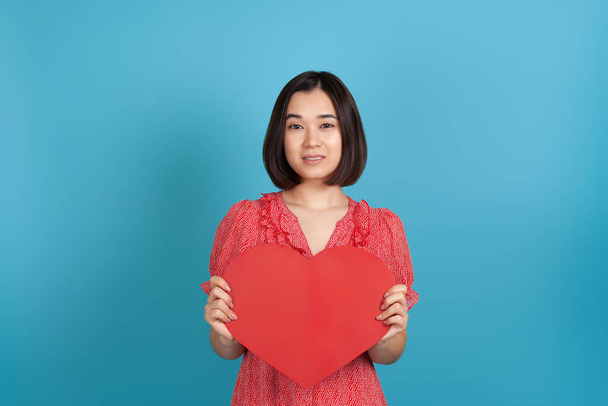 close-up smiling beautiful young Asian woman in a red dress holding a large red paper heart at her chest, isolated on a blue background - Фото, изображение