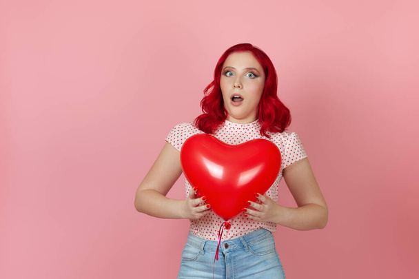 Close-up a surprised, shocked young woman with red hair holds a large flying red heart-shaped balloon isolated on a pink background - Photo, image