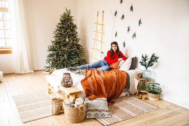 young beautiful girl in a red sweater and blue jeans sits on a bed in a cozy room decorated with Christmas decoration - Photo, Image