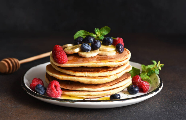 Homemade pancakes with banana, raspberries, blueberries and honey on a dark concrete background. - Foto, Imagen