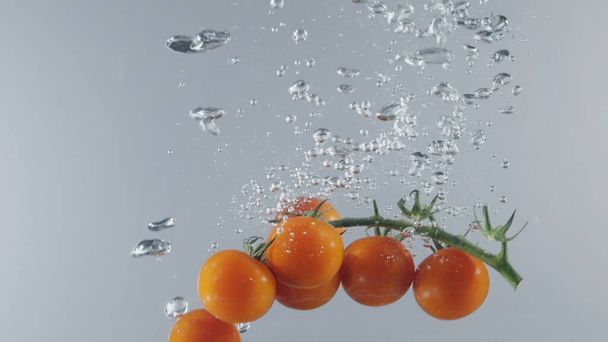 Гілка вишневих помідорів потрапляє в прозору воду на світло-сірому тлі стін. Дія. Свіжі органічні ягоди з зеленим стеблом в акваріумі, продуктовий магазин, здорова їжа
 - Фото, зображення
