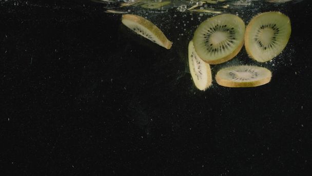 Kawałki owoców kiwi wpadają do wody pełnej bąbelków w zwolnionym tempie. Akcja. Zbliżenie ciętych owoców tropikalnych spadających pod wodą odizolowanych na czarnym tle. - Zdjęcie, obraz