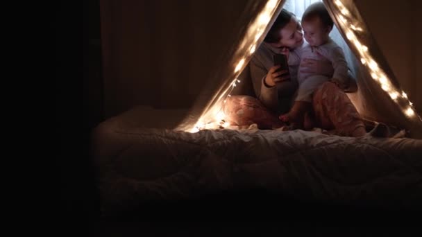 authentic cute mom with chubby babe infant girl in tent at home. young woman use smartphone with kid 6-12 months old sit in wigwam at night. Family, Childhood, Motherhood, Comfort and Safety concept. - Footage, Video