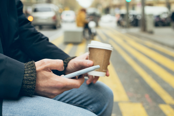Close up of man hands holding mobile phone, using internet while sitting on the bench and drinking take away cup of coffee. Chatting with friends or family, reading latest news and updates in the web. - Photo, Image