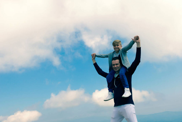Speelse vader houdt zoon op de schouders en heeft plezier op bergtop tegen de hemel. Kopieerruimte.  - Foto, afbeelding