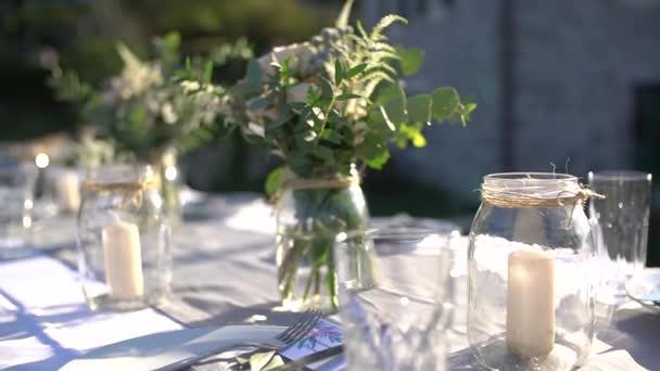 Stoły na bankiecie weselnym ze świecami w szklanych słoikach i bukietach róż, gałęzi astilby i eukaliptusa - Materiał filmowy, wideo