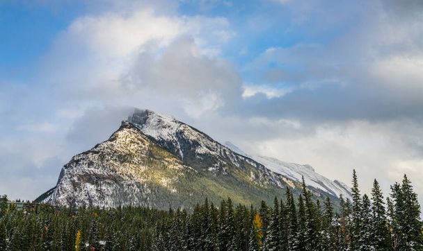 Zaśnieżony łańcuch górski Mount Rundle z zaśnieżonym lasem nad błękitnym niebem i białymi chmurami w zimowy słoneczny dzień. Park Narodowy Banff, Canadian Rockies. - Zdjęcie, obraz