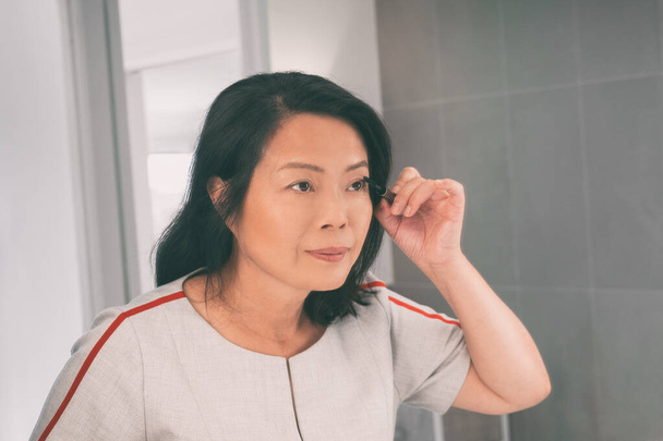 Makeup Asian mature woman putting mascara looking in make-up mirror getting ready in the morning. Middle aged chinese lady in her 50s - Foto, Imagen