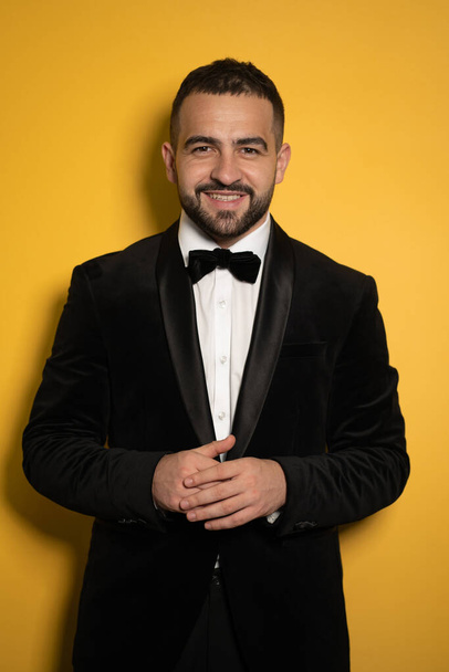 Smiling handsome man in black tuxedo with hands folded looking on camera, work profession lifestyle. Handsome young smiling caucasian man isolated on yellow background. Male fashion concept - Foto, Imagen